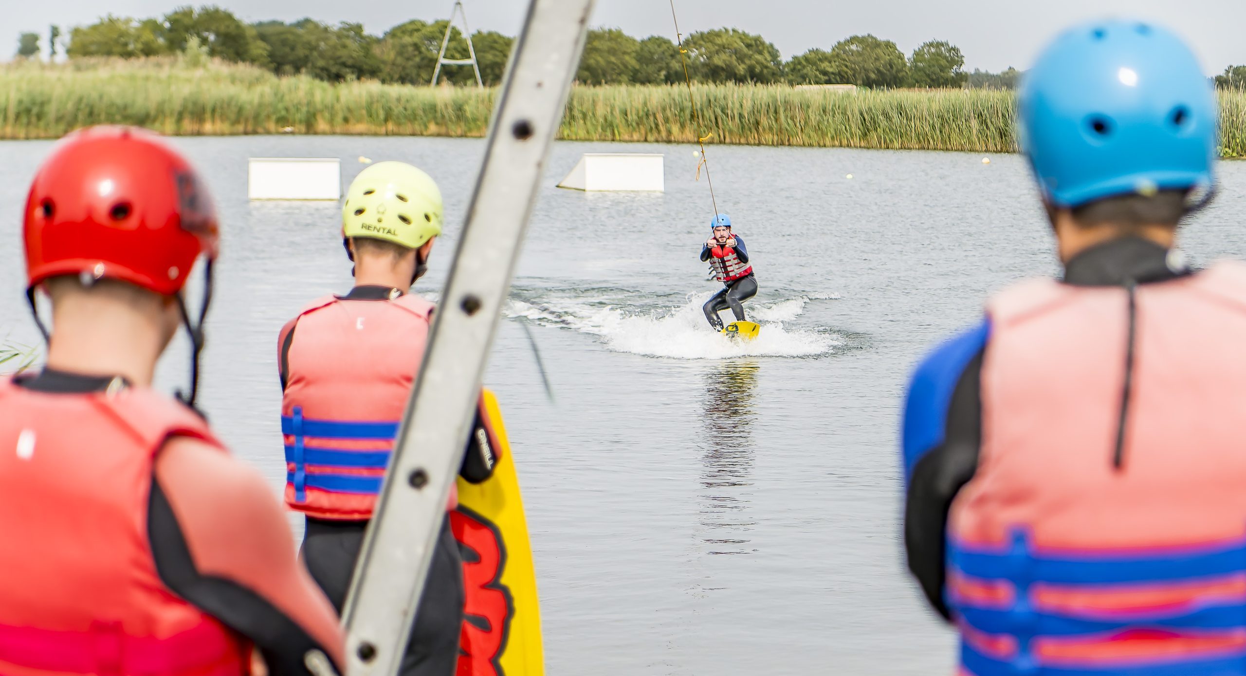 Observers in red and blue life jackets stand on a dock watching a wakeboarder glide across the lake, spraying water behind.
